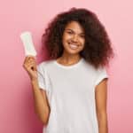 Glad attractive curly female model holds thin sanitary pad, uses feminine intimate product during menstruation or periods, cares about hygiene, wears casual t shirt, models over pink background
