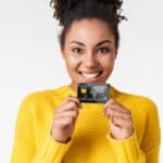 Melhores Cartões de Crédito Sem Anuidade Disponíveis no Brasil African excited emotional happy woman posing isolated over white wall background holding credit card.