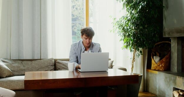 A person using a laptop while sitting at a desk in a home office. There is a notepad and pen next to the laptop, and the screen shows a website with financial charts and information about unemployment benefits.