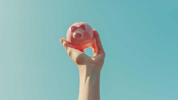 A close-up shot of a hand placing a coin into a piggy bank shaped like a school bus. The background is softly blurred, showing textbooks and a globe, symbolizing saving for education. The focus is on the hand and the coin being deposited, emphasizing the act of saving.